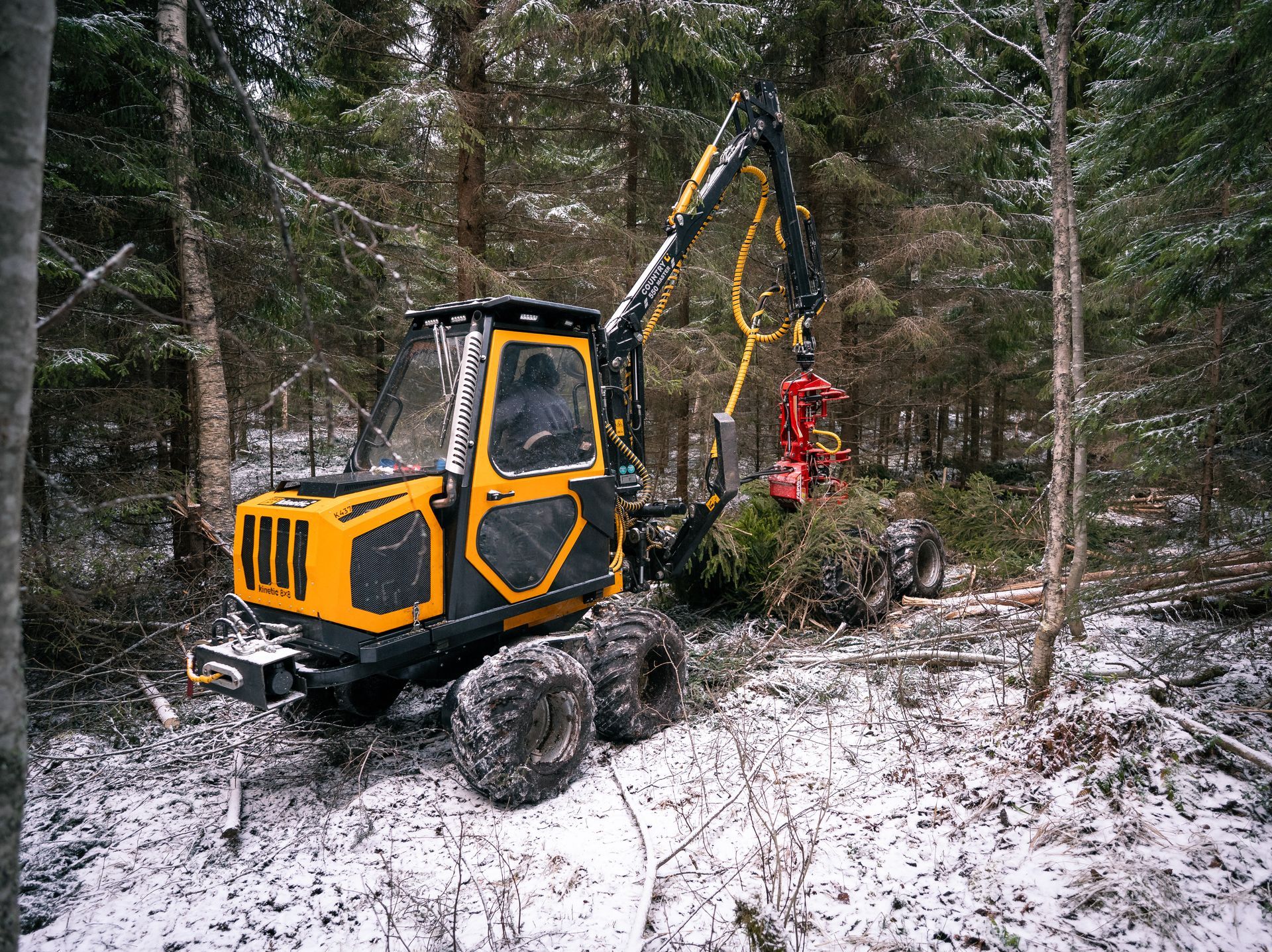 Keltainen metsäkone lumisessa metsässä kaatamassa puita.