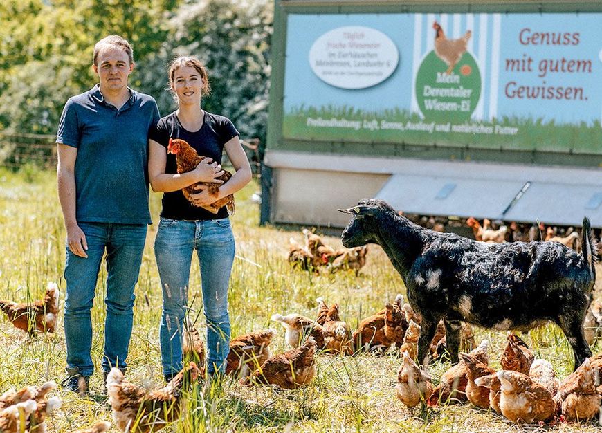 Ein Mann und eine Frau stehen mit Hühnern und einer Ziege auf einem Feld.