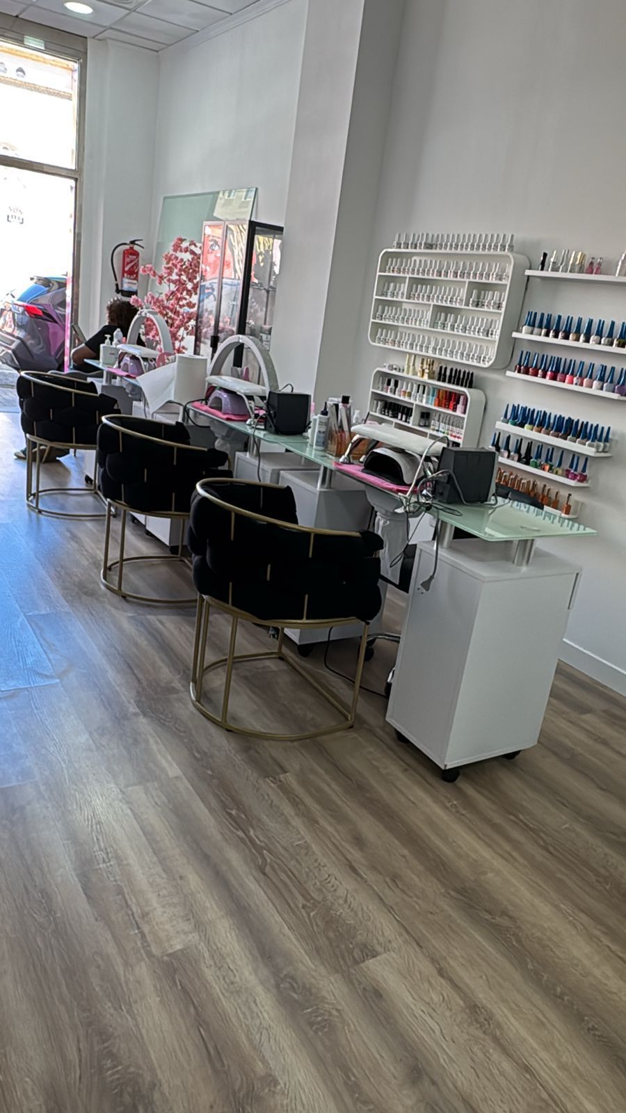 Interior del salón con mesas de manicura, estantes para esmaltes de uñas y grandes ventanales sobre suelo de madera.