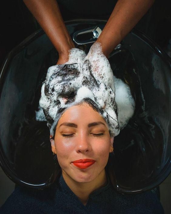 Mujer recostada en un lavacabezas de peluquería con el cabello cubierto de espuma blanca y los ojos cerrados.