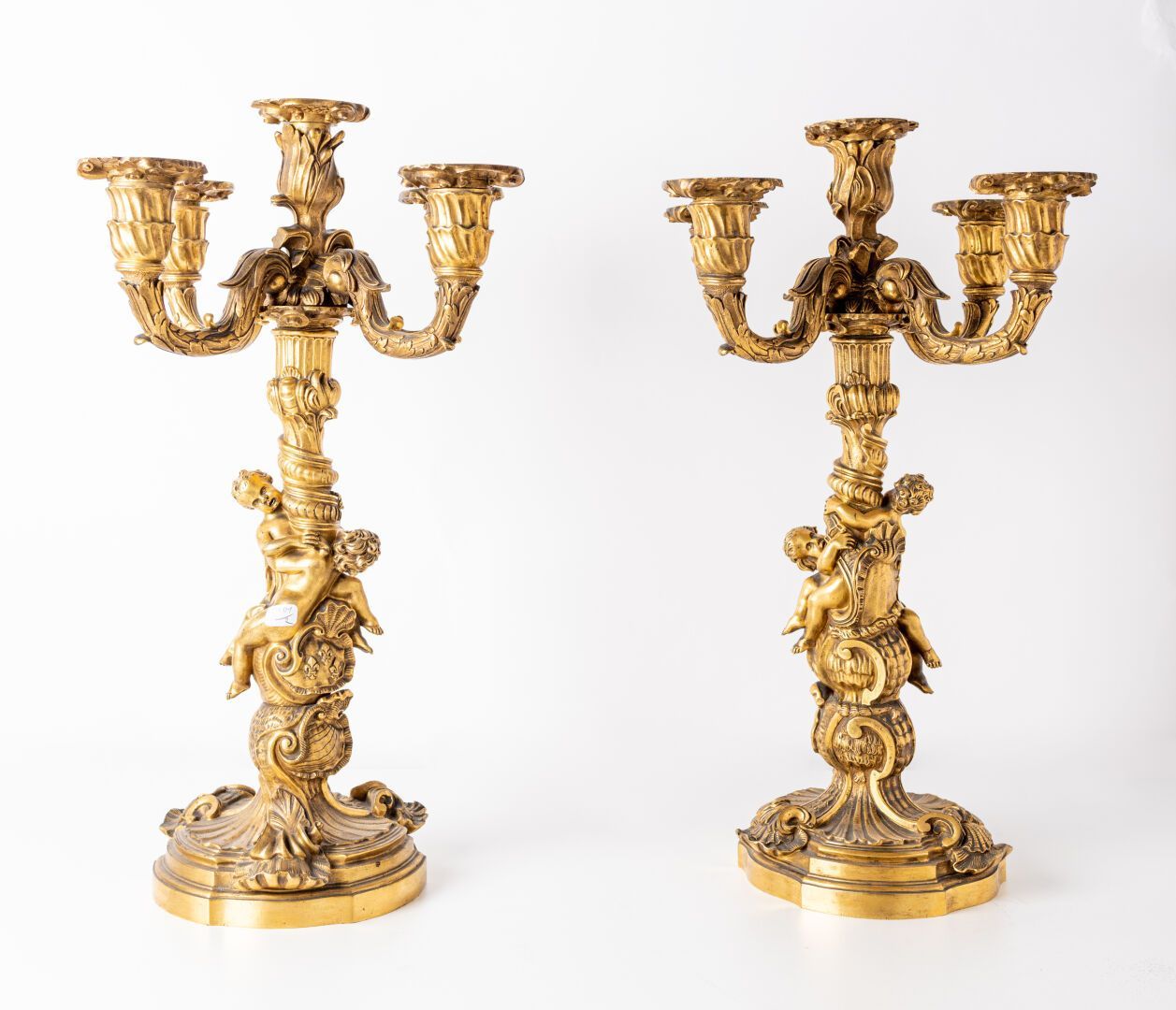 Deux candélabres dorés ornés avec trois bougeoirs, des chérubins et une base décorative sur fond blanc.