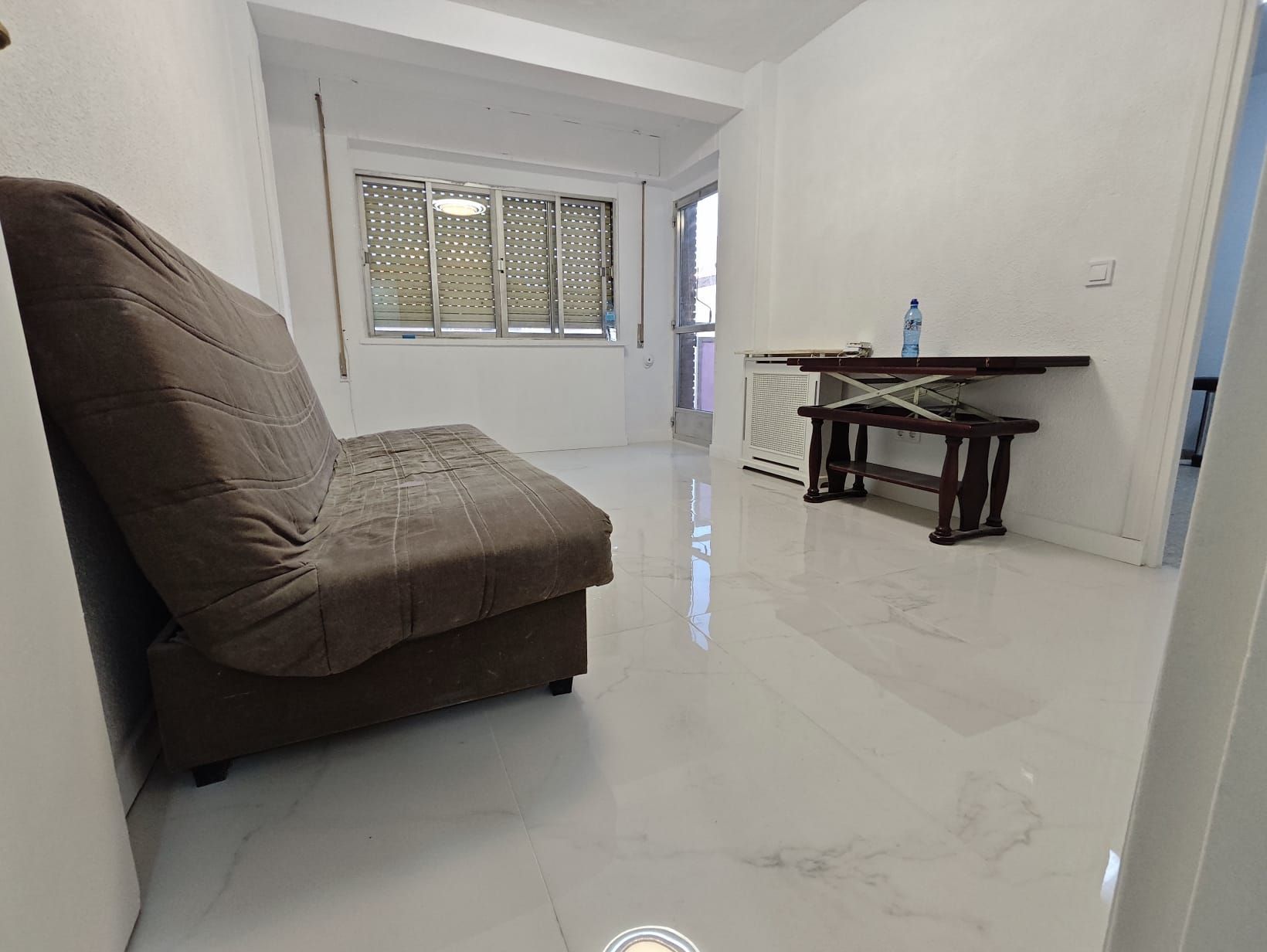 Una pequeña sala de estar con un futón marrón, una mesa de madera y una ventana con persianas. Paredes y suelo blancos.