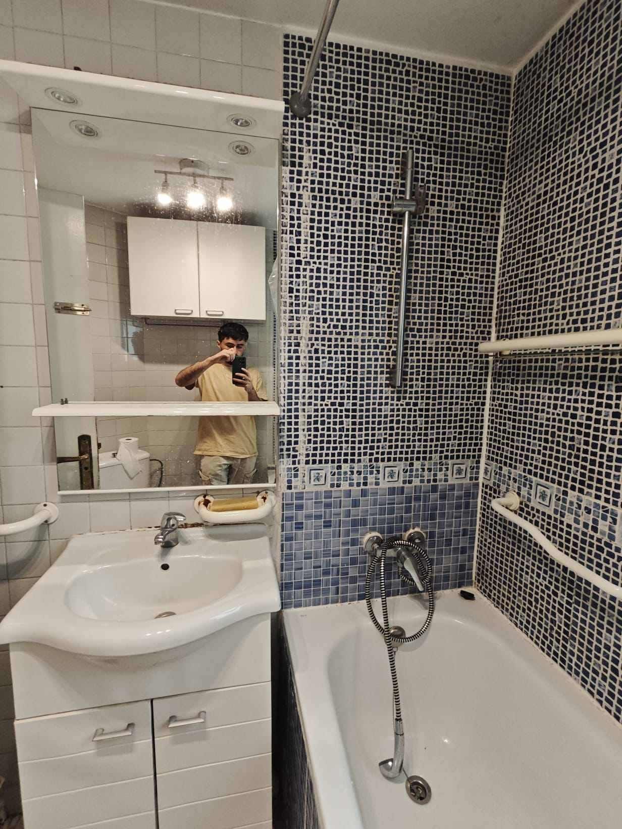 Baño con lavabo blanco, bañera y azulejos azules. Una persona se refleja en el espejo tomándose una foto.