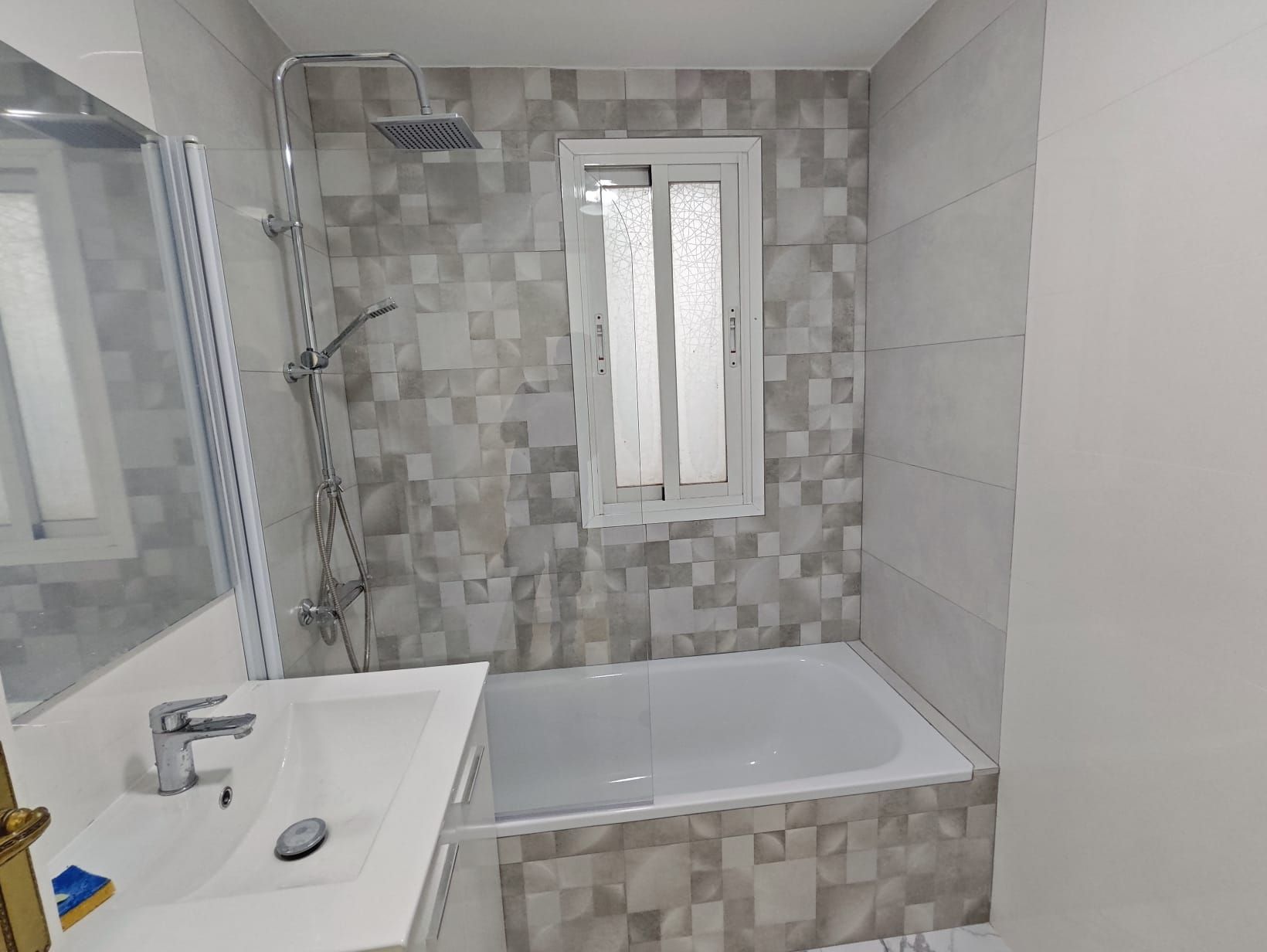 Un baño con bañera, ducha, lavabo y ventana. Las paredes están revestidas con un mosaico gris y blanco.
