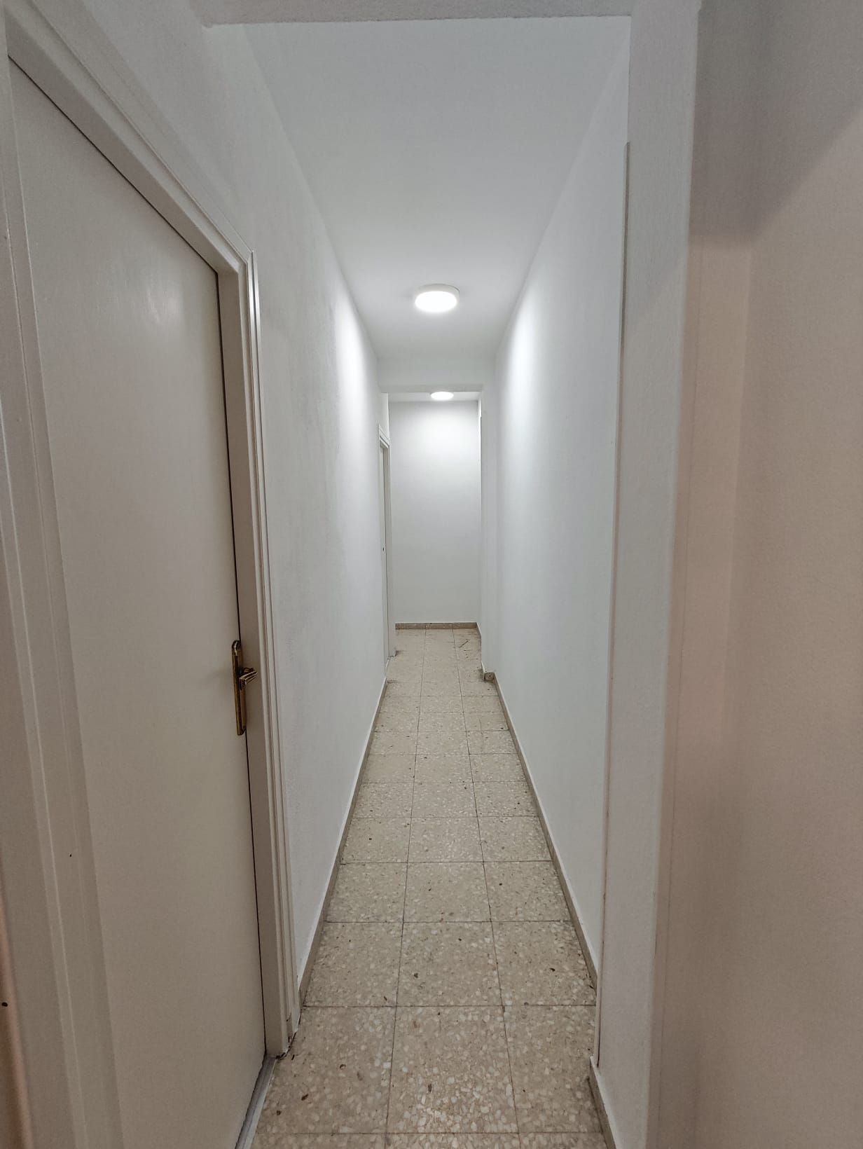 Pasillo estrecho con paredes blancas, una puerta cerrada a la izquierda e iluminación empotrada. El suelo es de baldosas.