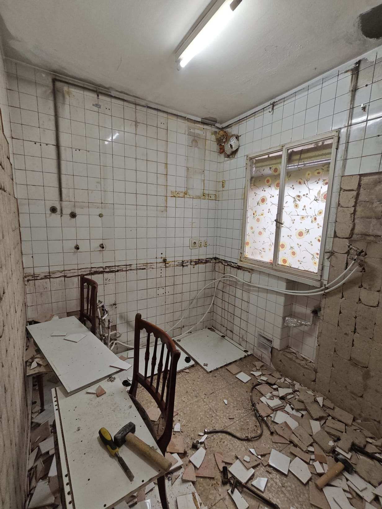 Una cocina en reforma, con paredes expuestas, azulejos rotos y una ventana. Hay una mesa y sillas en la habitación.