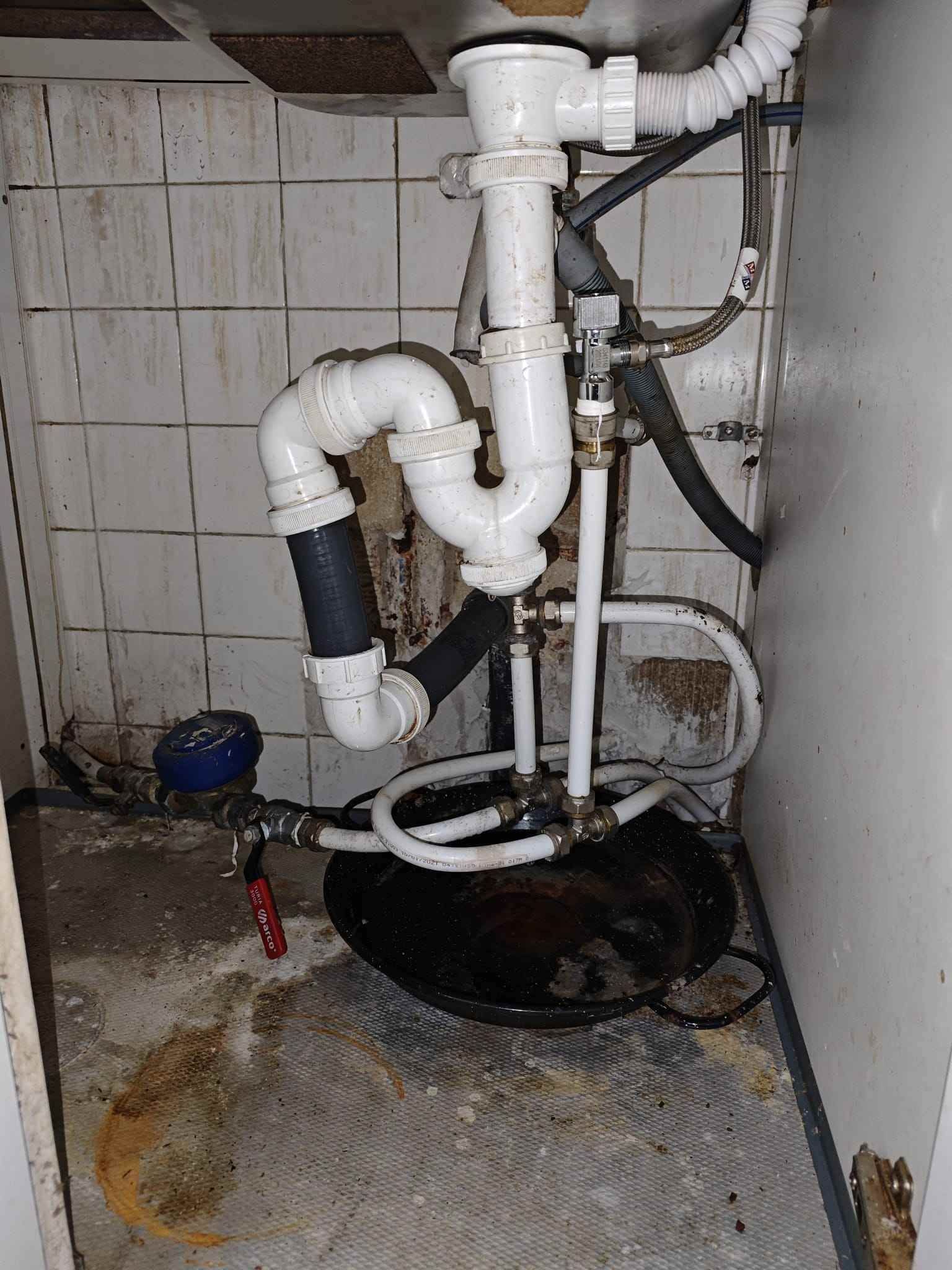 Fontanería debajo del fregadero: Tuberías blancas forman un sifón. Se ven manchas de agua en el suelo circundante.