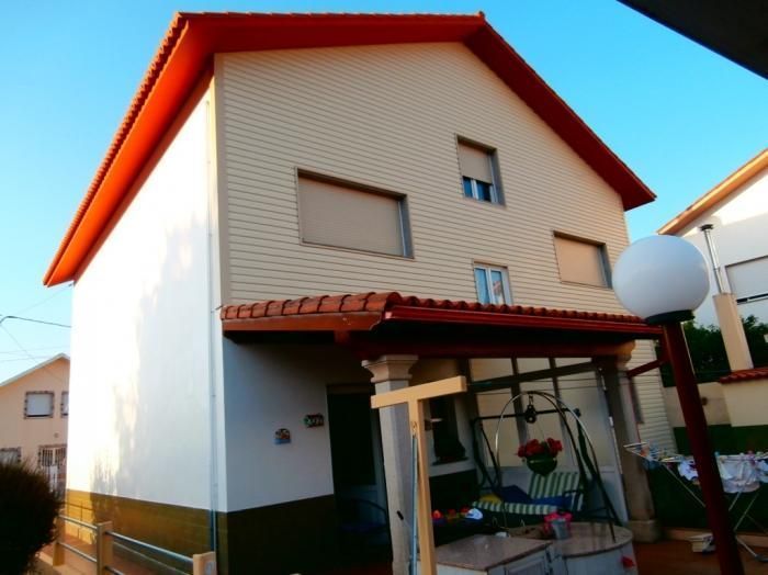 Casa de dos pisos con revestimiento de color canela, techo rojo y toldo, bajo un cielo azul claro.