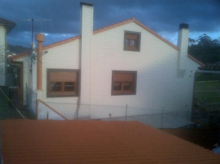 Casa blanca con tejado de color naranja, dos chimeneas y dos ventanas con contraventanas marrones, bajo un cielo nublado.