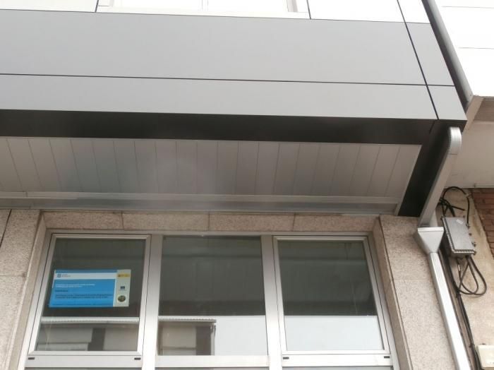 Fachada de edificio con paneles grises y blancos sobre las ventanas; se ven un canalón y una caja eléctrica.