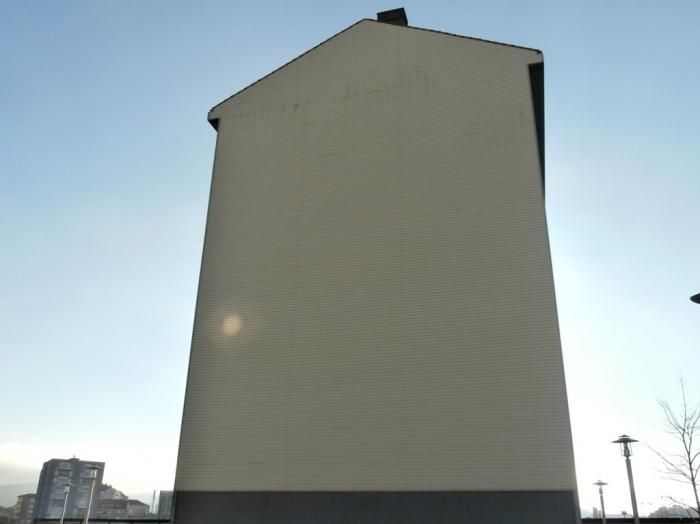 Edificio alto de color crema con base gris oscuro sobre un cielo azul claro. La luz del sol brilla cerca del centro.