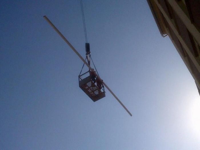 Trabajadores en una cesta suspendida, sujeta a una grúa, sujetando una viga larga contra la fachada de un edificio. Cielo azul despejado.