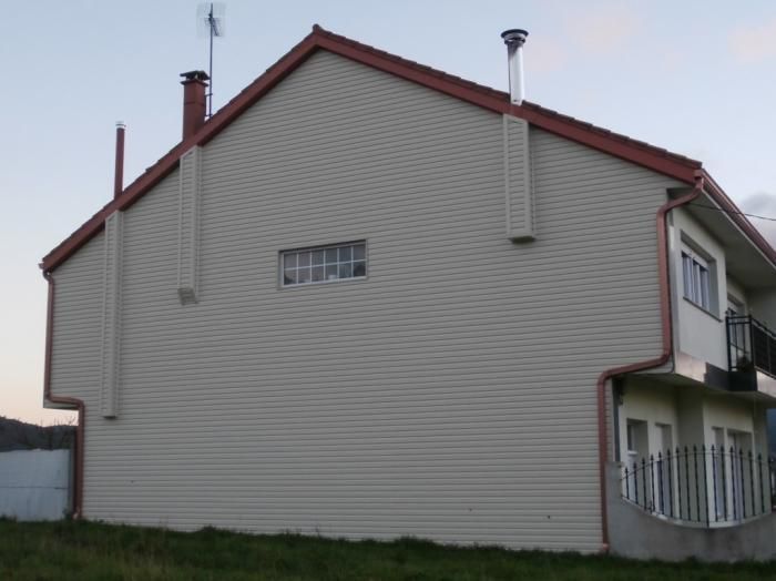 Casa beige de dos plantas con tejado rojo. Dos chimeneas metálicas se extienden hacia arriba. Césped en primer plano, cielo al fondo.