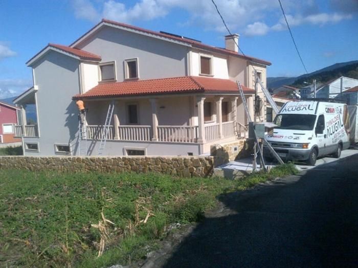Casa de dos pisos con techo de tejas rojas, paredes beige y porche. Un trabajador subido a una escalera y una camioneta estacionada cerca.