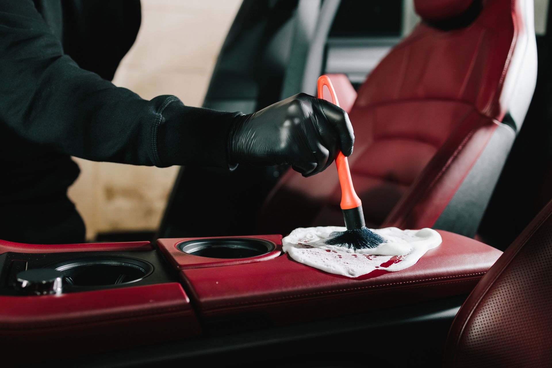 Manos enguantadas limpiando la consola roja de un coche con un cepillo y un paño.