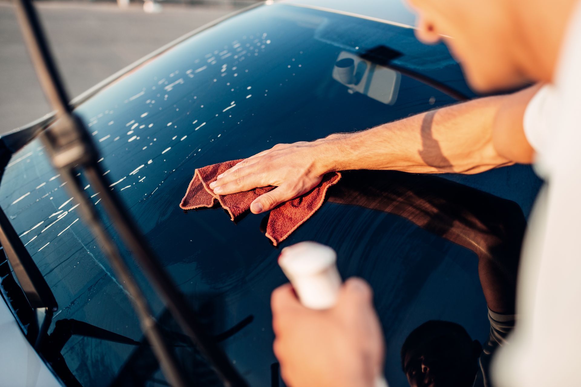 Persona limpiando el parabrisas de un coche con un paño rojo y una botella con pulverizador.