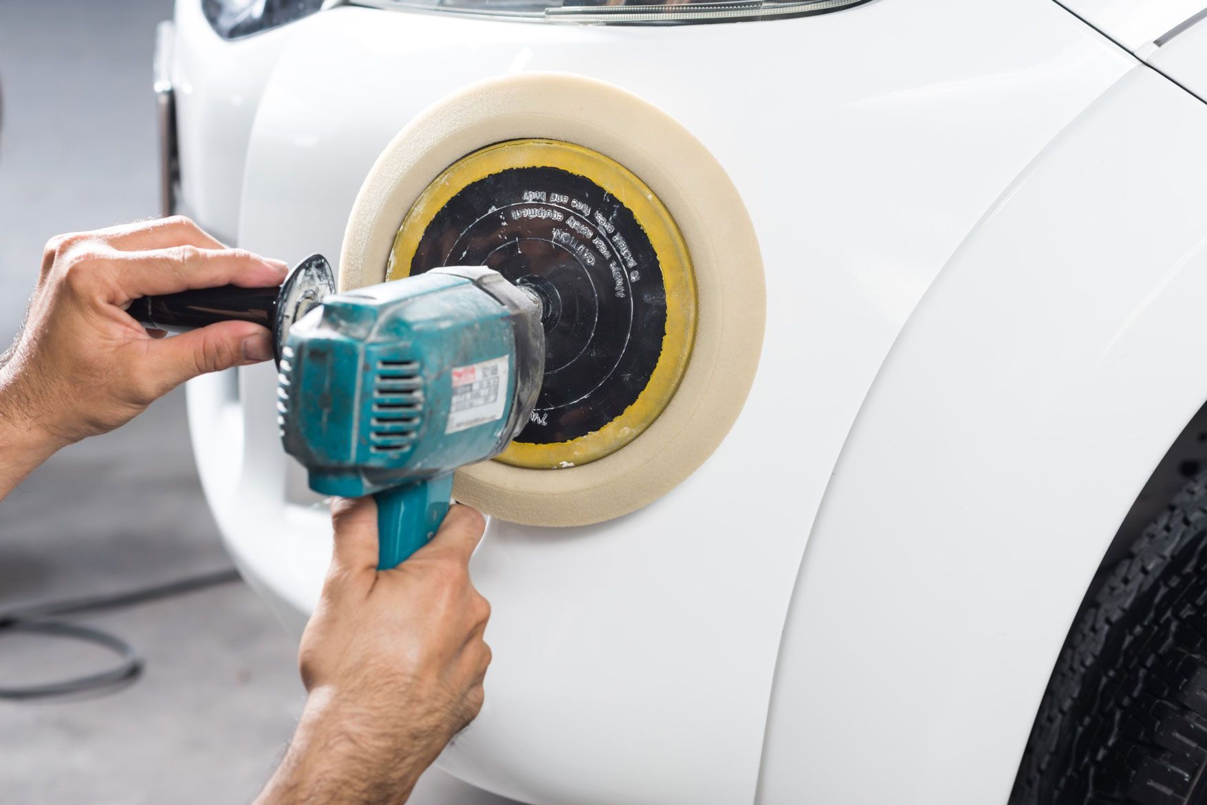 Persona utilizando una pulidora para pulir el panel de la carrocería de un coche blanco; la herramienta es azul y negra.