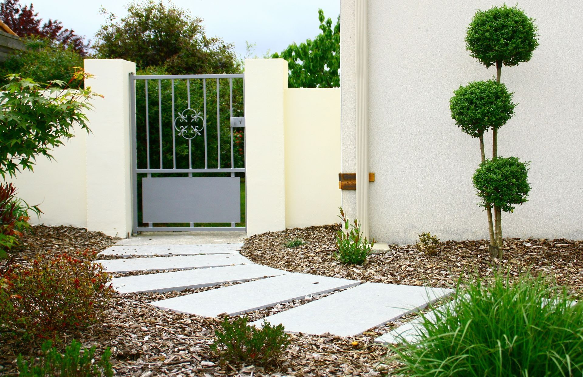 Un chemin de pierre mène à un portail métallique au sein d'un jardin moderne ; des arbustes taillés longent un mur en stuc.