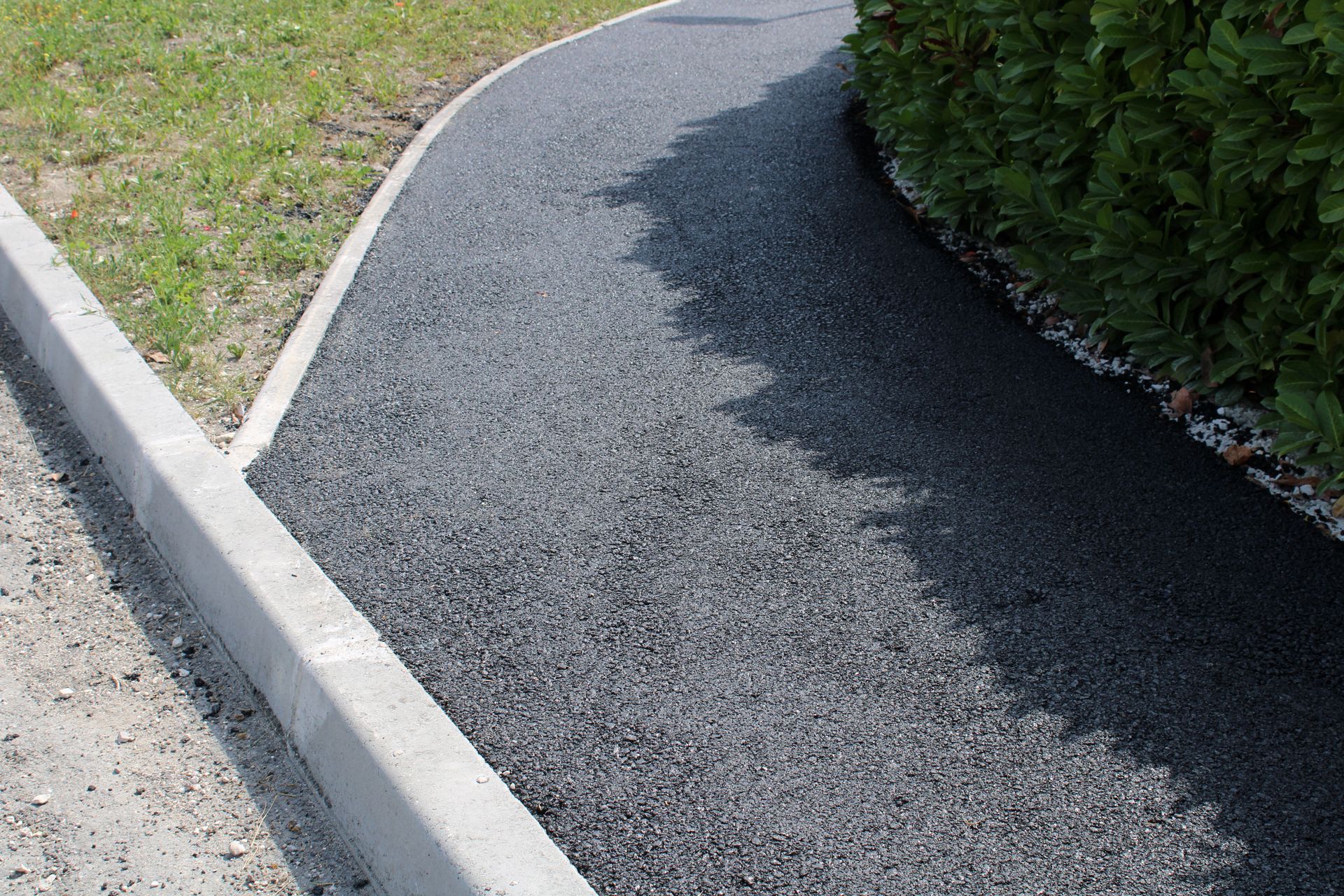 Un chemin de gravier serpente le long d'une haie verte et d'une pelouse, avec un trottoir en béton d'un côté.