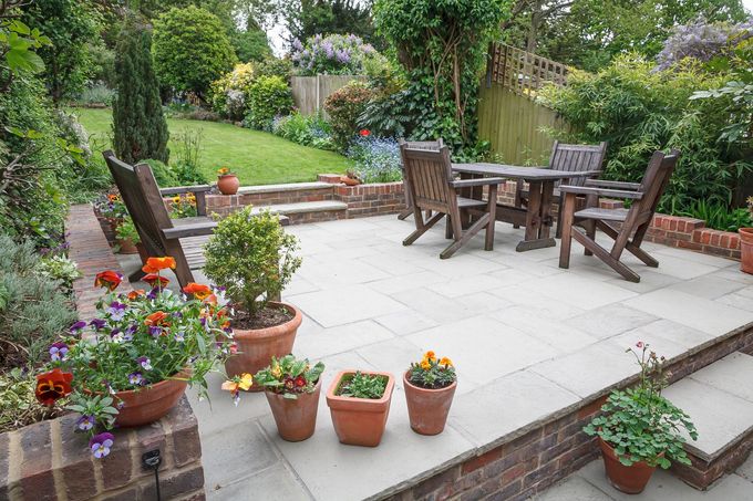 Terrasse en pierre avec table et chaises en bois, entourée de fleurs en pot et de verdure dans un cadre de jardin.
