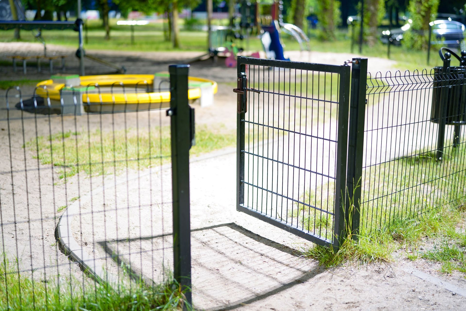 Portail métallique noir ouvert dans une aire de jeux, donnant sur un bac à sable et des équipements de jeux.