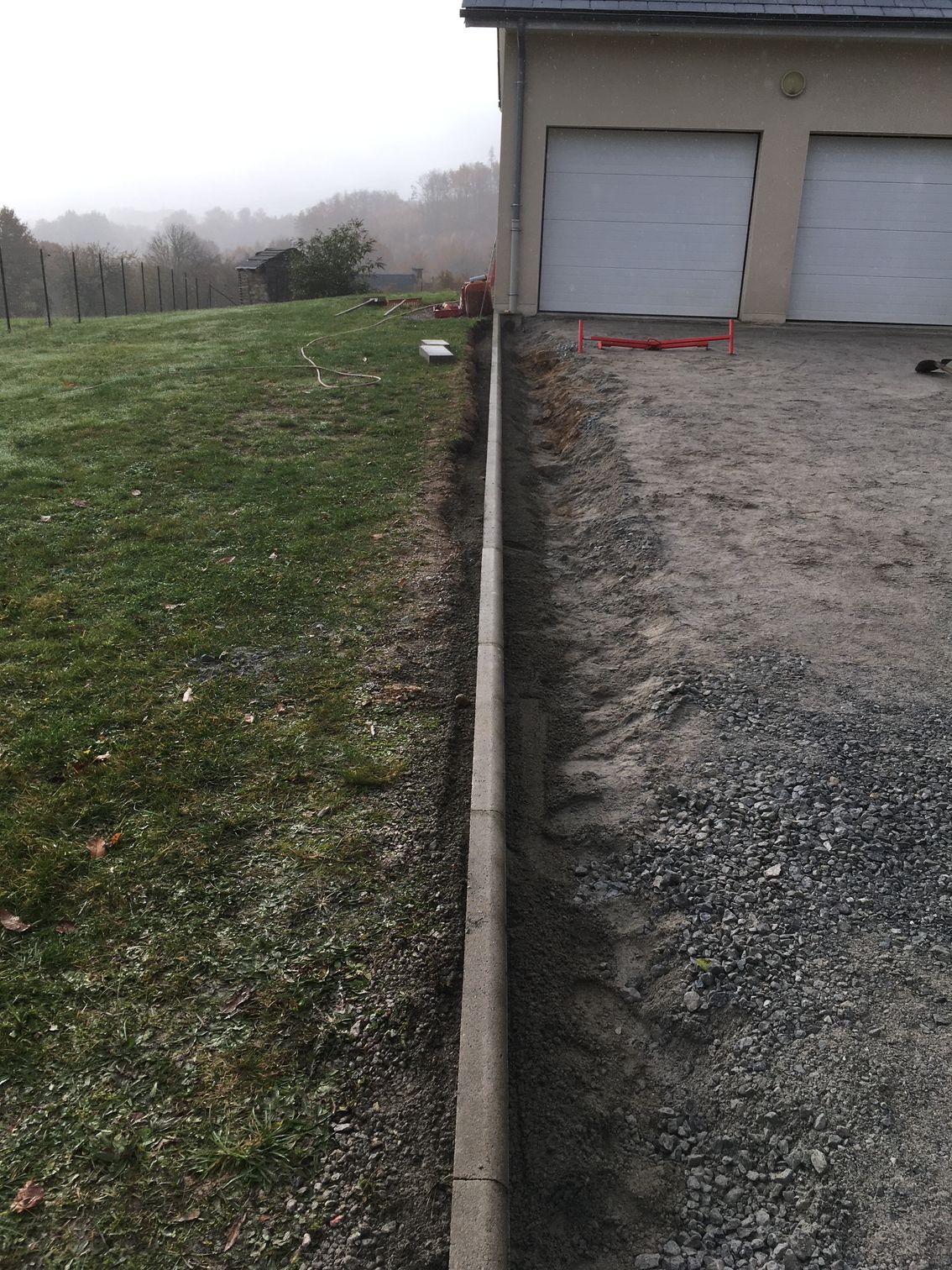Pelouse et allée de gravier séparées par une bordure en béton. Maison avec portes de garage en arrière-plan. Ciel couvert.