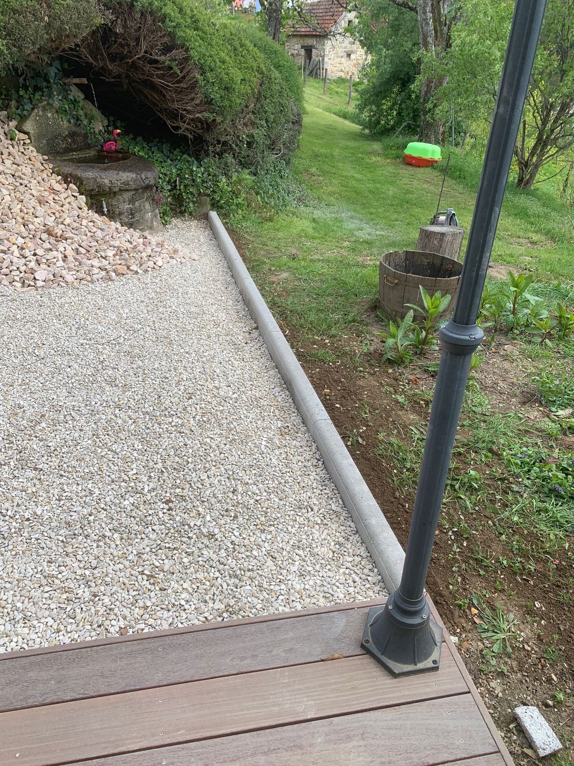 Terrasse en gravier jouxtant une zone gazonnée, avec un poteau métallique au premier plan et une haie à l'arrière-plan.