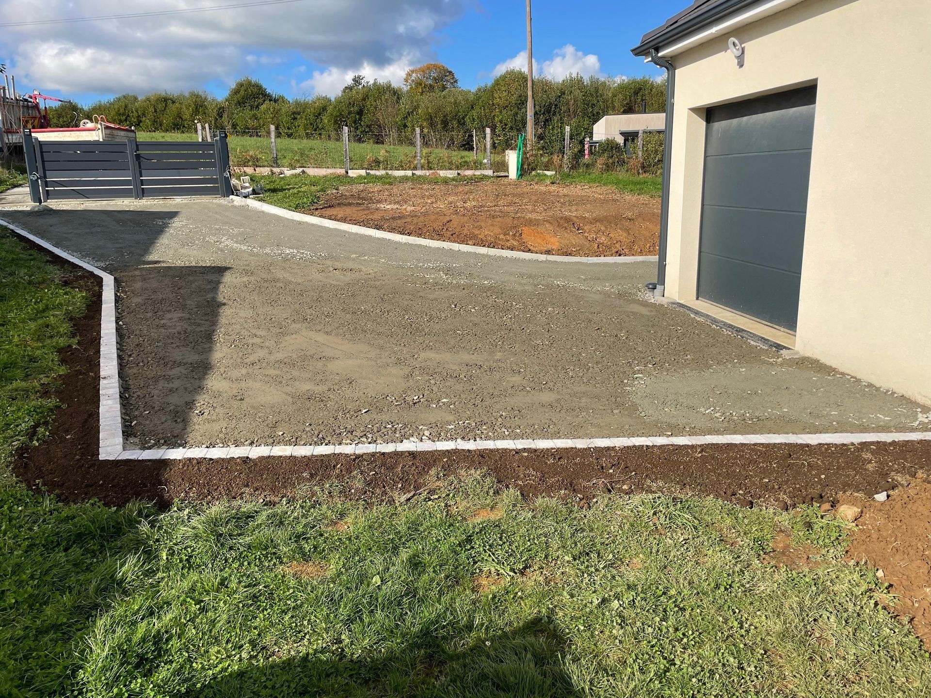 Allée de gravier bordée de galets argentés, jouxtant un bâtiment beige avec une porte de garage grise fermée.