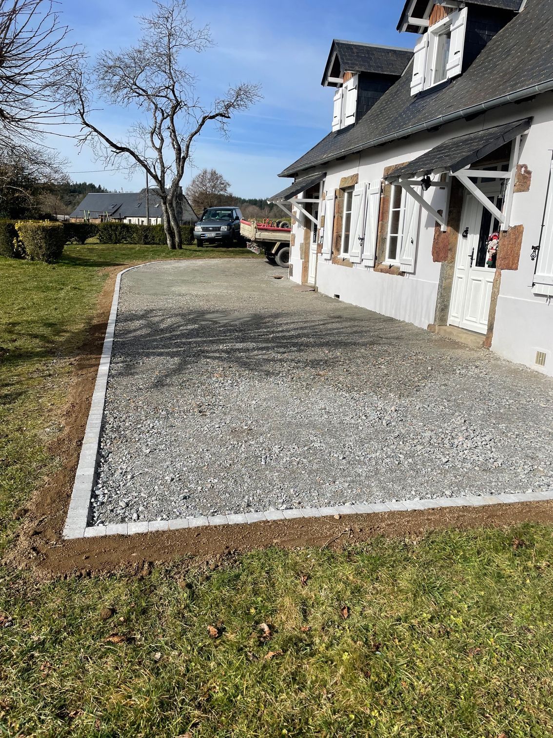 Terrasse en gravier bordée d'une clôture jouxtant une maison blanche à volets, dans un jardin gazonné. Un camion est stationné à l'arrière-plan.