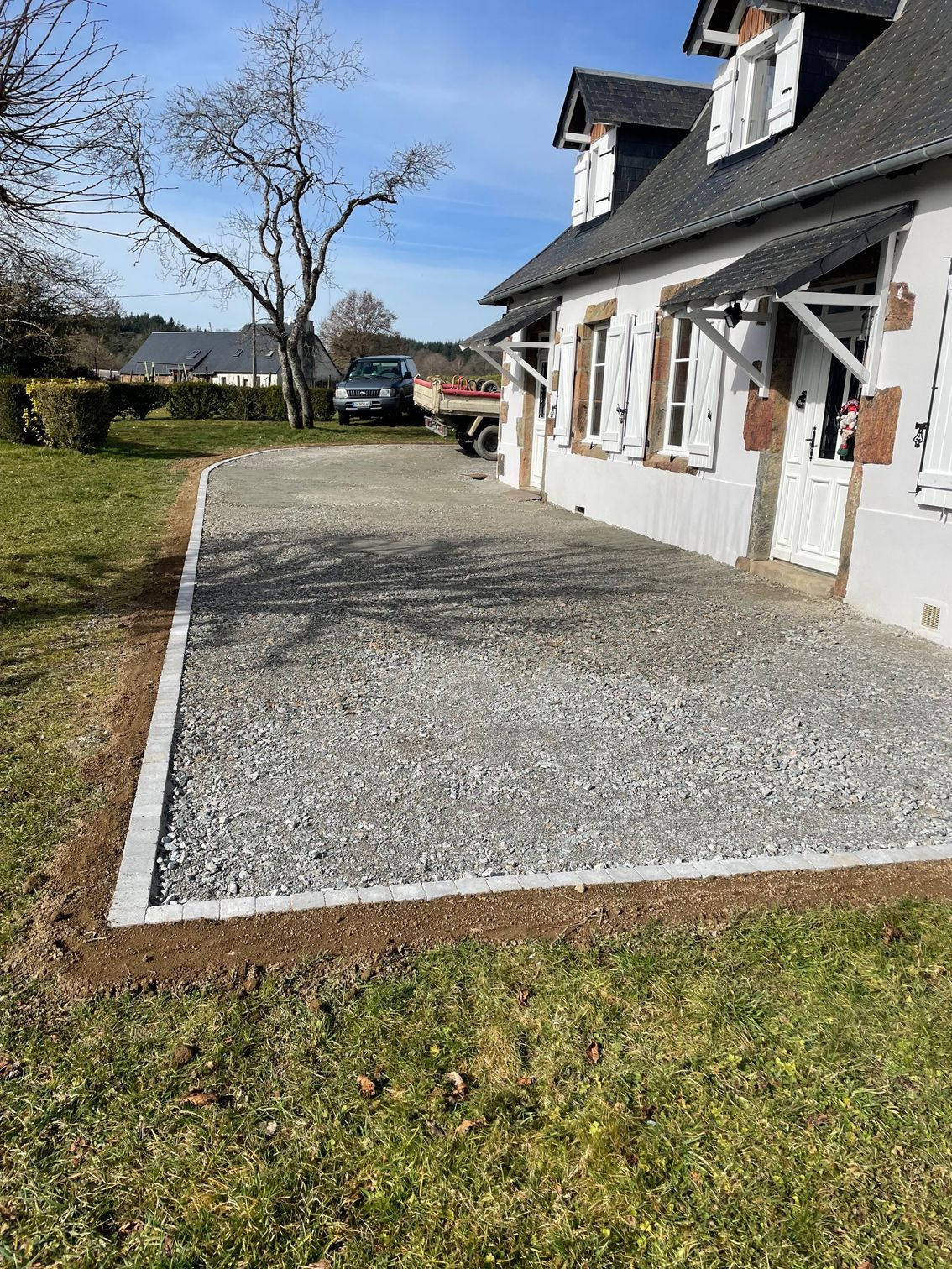 Allée de gravier bordée de pavés, à côté d'une maison blanche à volets, sur une pelouse sous un ciel bleu.