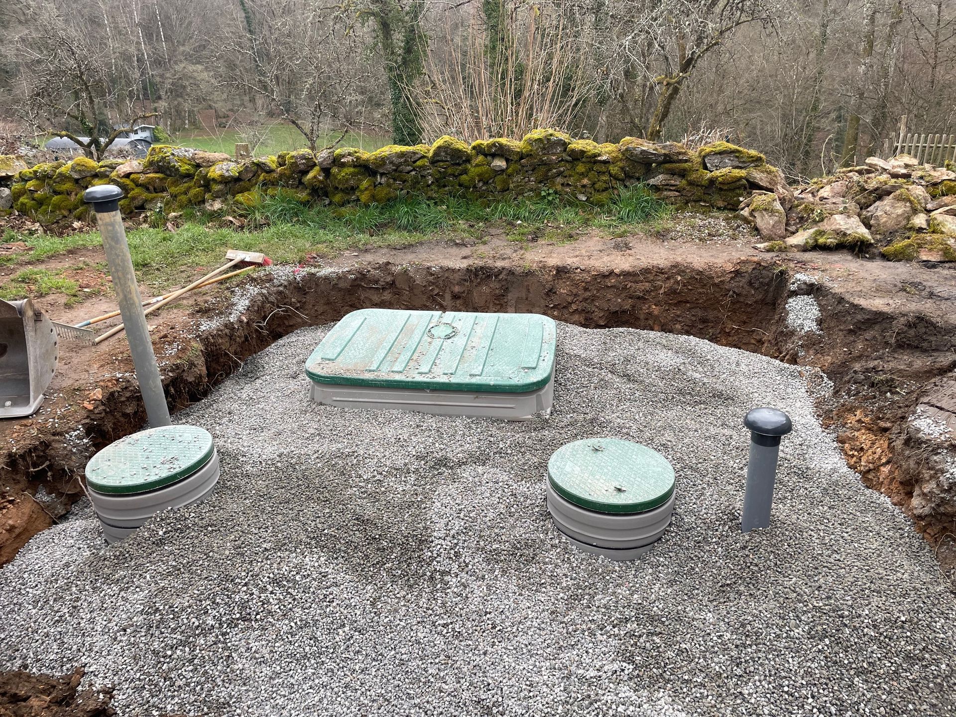 Une excavation remplie de gravier avec un couvercle de fosse septique vert et deux points d'accès circulaires, un tuyau gris et de la verdure en arrière-plan.