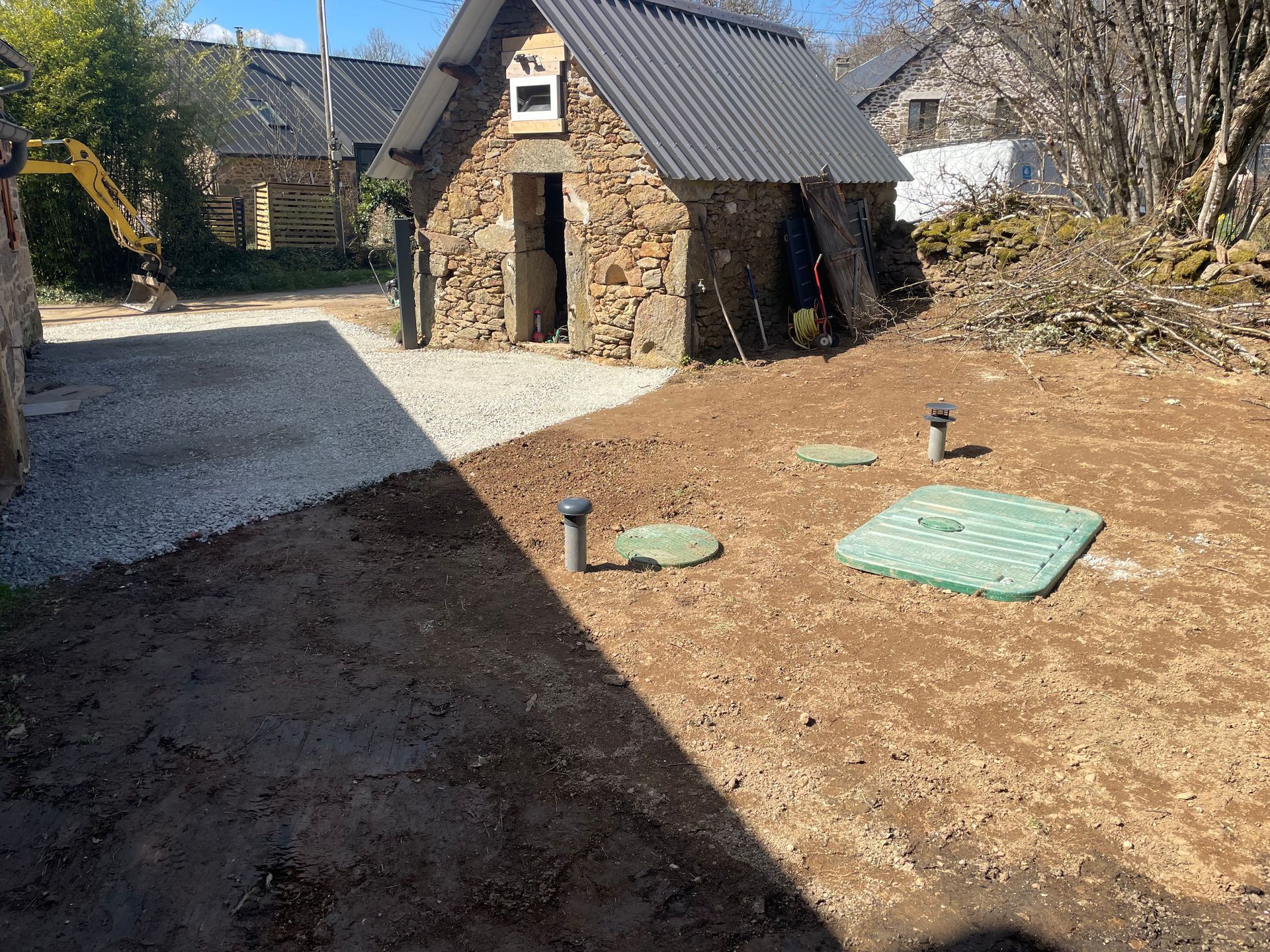 Un bâtiment en pierre avec un toit en tôle ondulée se trouve dans une cour. Un chemin de gravier se trouve à gauche. On aperçoit les couvercles verts de la fosse septique.