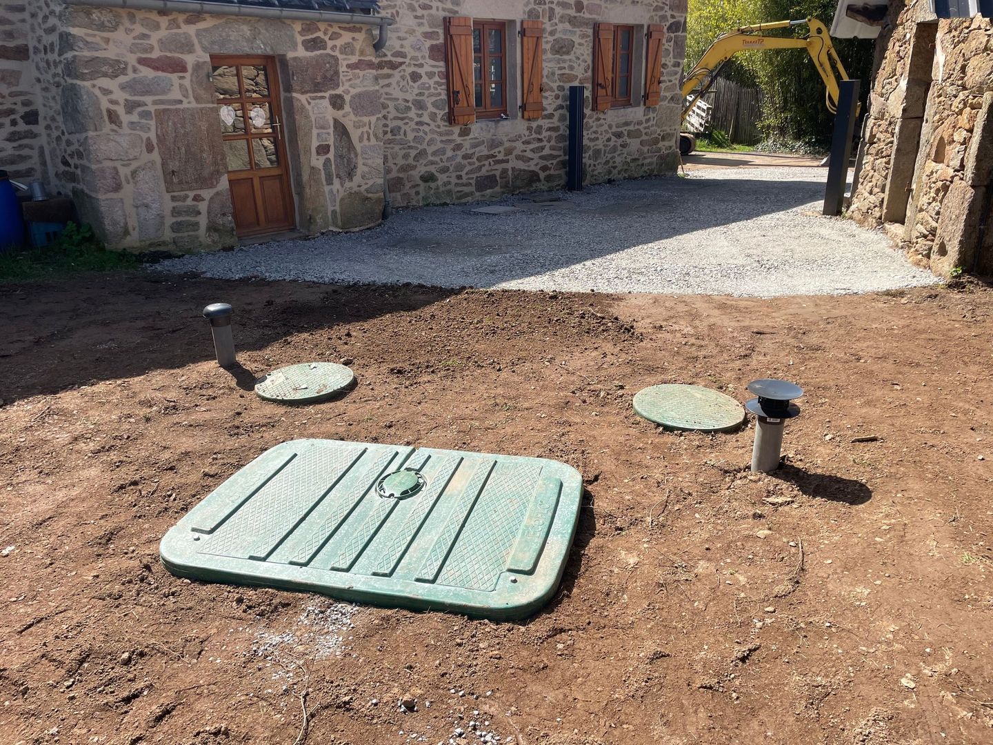 Couvercles de fosses septiques vertes dans une cour en terre battue près d'une maison en pierre.
