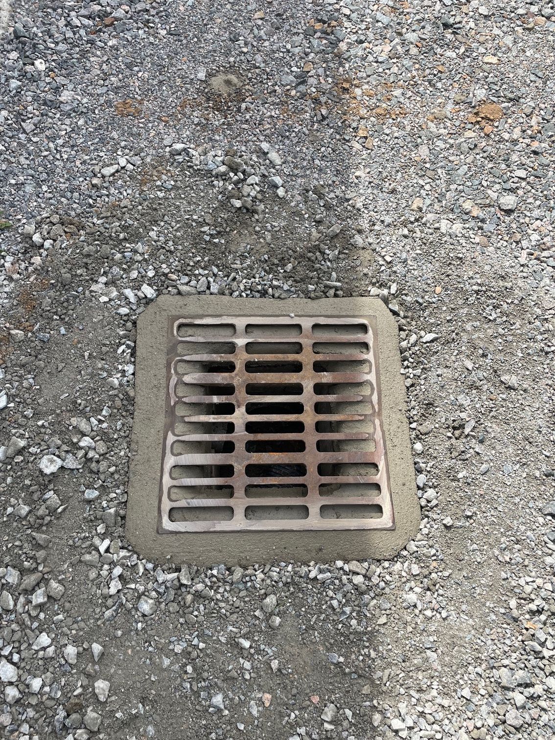 Grille métallique carrée scellée dans du béton et entourée de gravier.
