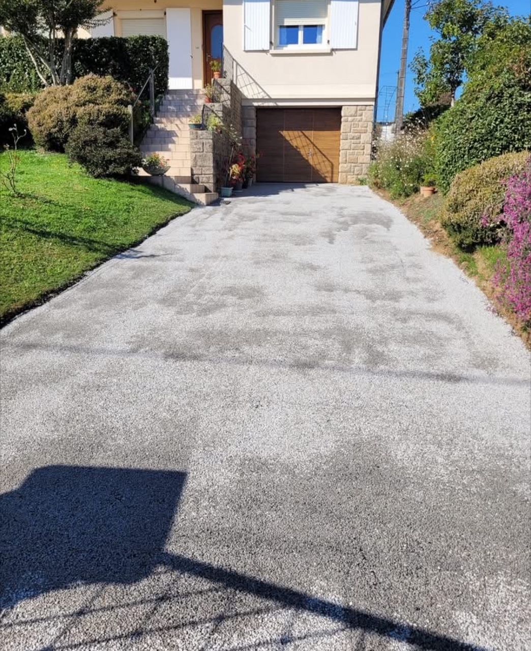 Une allée en béton monte vers une maison avec garage et porte d'entrée. De la pelouse et des buissons bordent la maison.