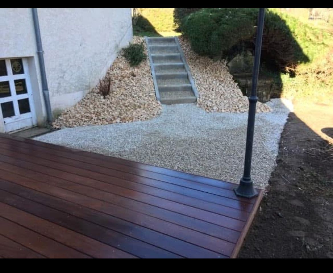 Terrasse en bois longeant un chemin de gravier gris clair et un escalier menant au sommet d'une petite colline.