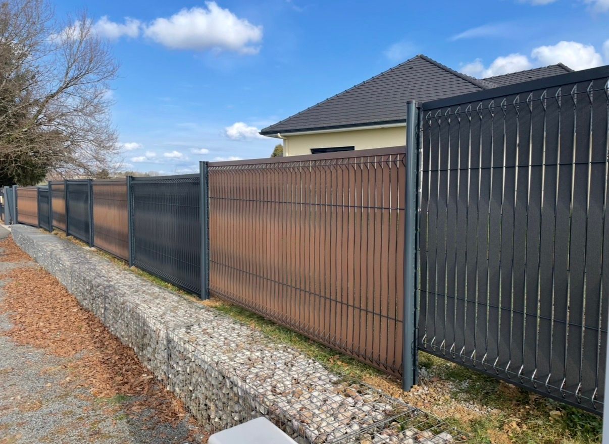 Clôture d'intimité brune et noire le long d'une allée de gravier devant une maison, sous un ciel bleu.