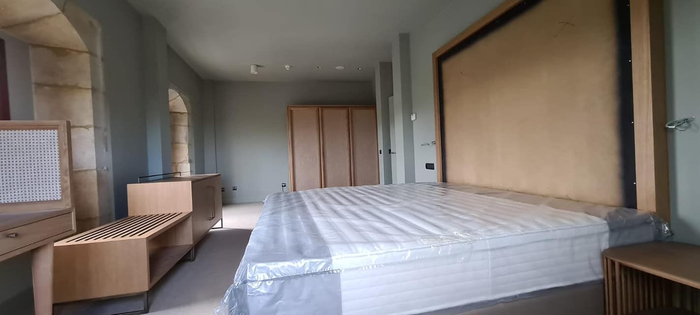 Dormitorio con cama revestida en plástico, amplio cabecero, muebles color marrón claro y paredes color gris claro.