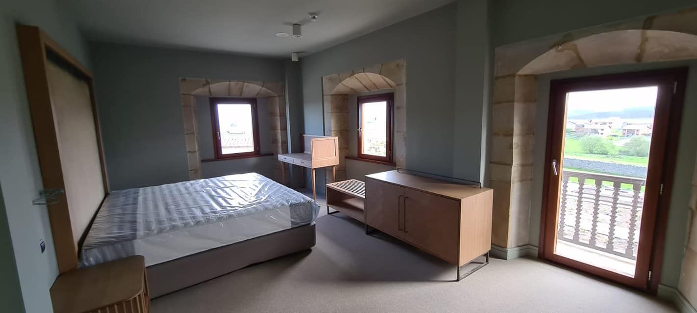 Dormitorio con cama revestida de plástico, dos ventanas y puerta a un balcón con arco de piedra.