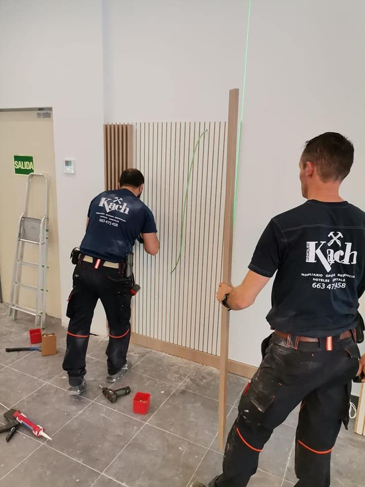 Dos carpinteros instalan paneles de madera en la pared utilizando un nivel láser en un espacio interior.