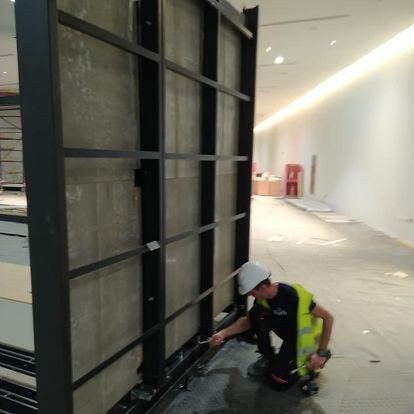 Un obrero ajusta una pared con estructura de acero en un espacio interior luminoso. Lleva casco y chaleco de seguridad.