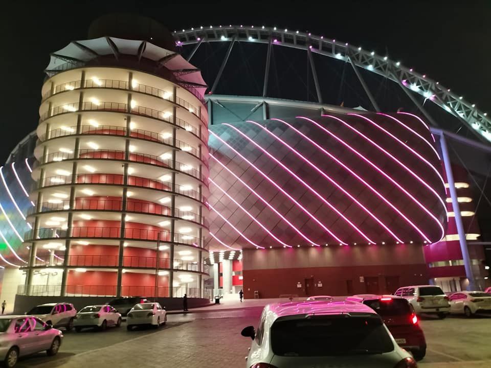 Estadio de noche con luces de neón rosas. Hay coches aparcados frente a un aparcamiento de varias plantas.