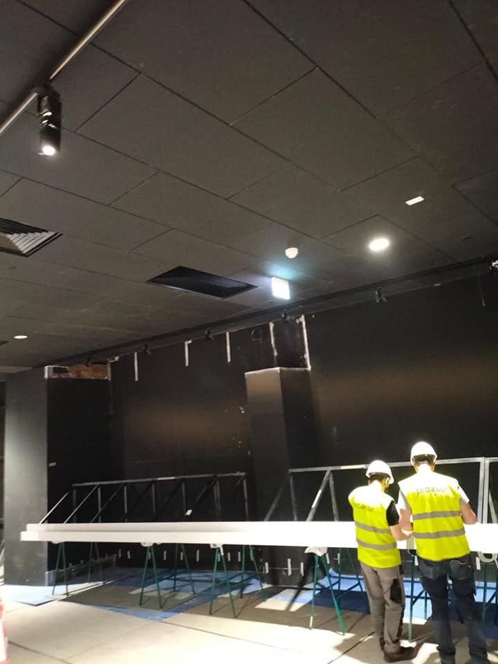 Trabajadores con chalecos salvavidas cerca de una pared negra con un escenario en construcción. Techo negro y focos.