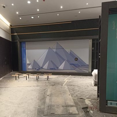 Interior con mural piramidal y marcos de exhibición. Gris y azul. Materiales de construcción visibles.
