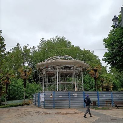 Quiosco de música en construcción, rodeado de andamios y una barrera azul, con una persona caminando.