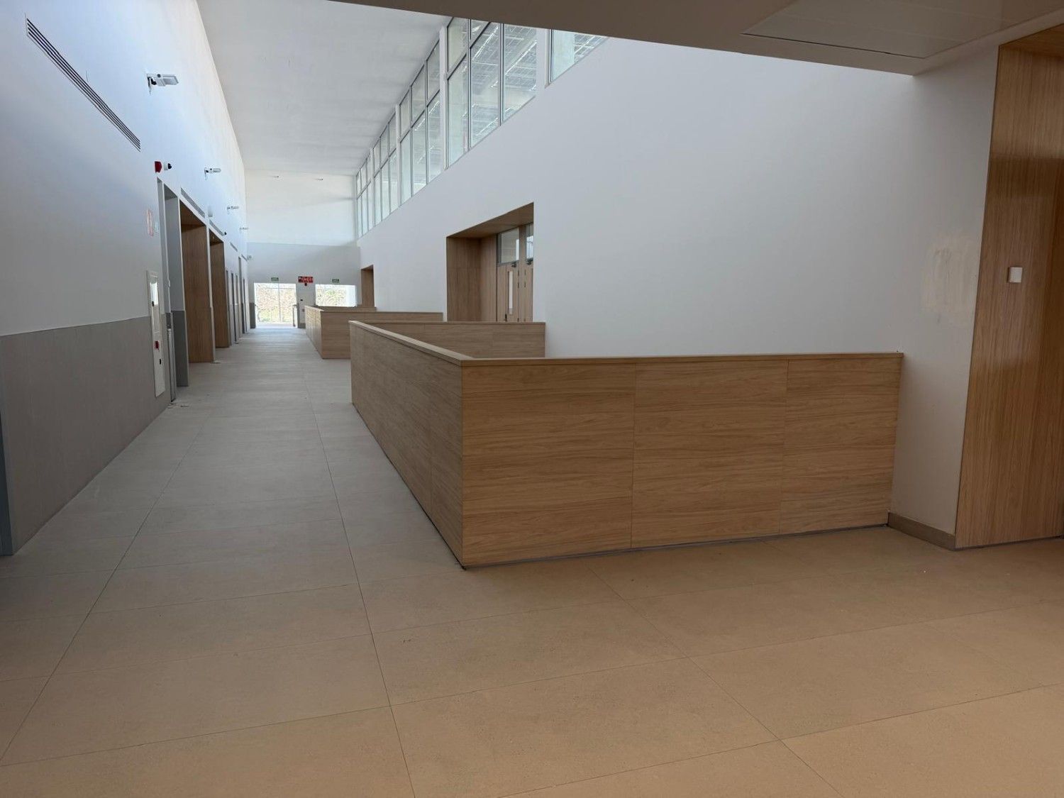 Pasillo largo y vacío con mostrador de recepción de madera de color claro, paredes blancas y ventanas altas.