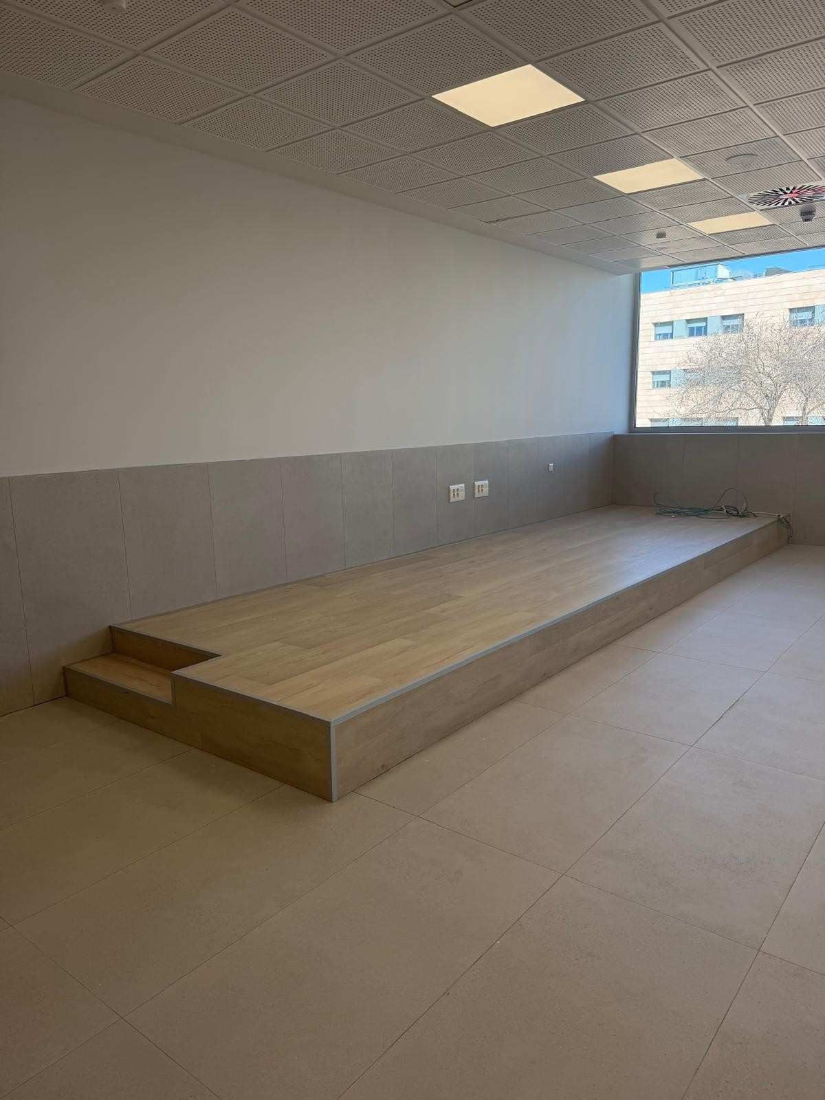 Tarima elevada de madera en habitación con gran ventanal, colores neutros y suelo de baldosas.