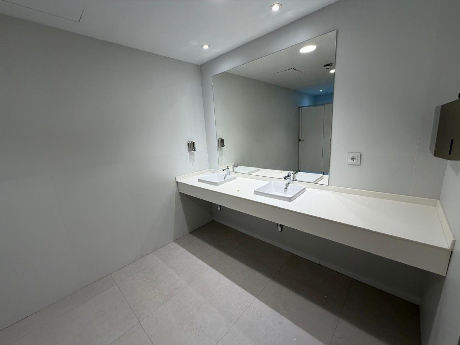 Baño público limpio y moderno con dos lavabos, espejo grande y dispensador de toallas de papel.