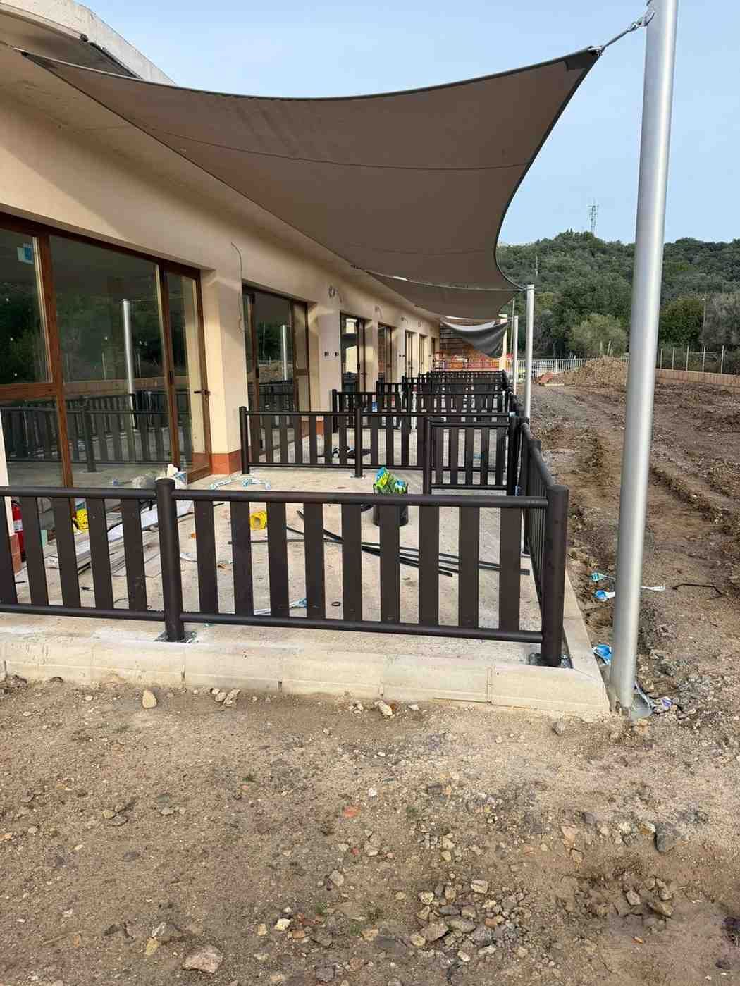 Un exterior de edificio con un patio sombreado, una cerca marrón y un suelo de tierra.