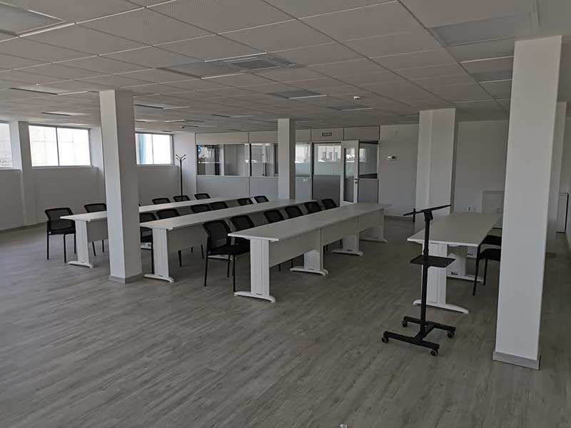 Aula vacía con largas mesas blancas, sillas negras y columnas blancas.